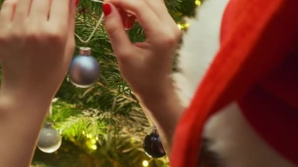 Kobieta Czerwonym Kapeluszu Mikołaja Dekoruje Drzewo Zabawką Piłkę Przeciwko Girlandzie — Wideo stockowe