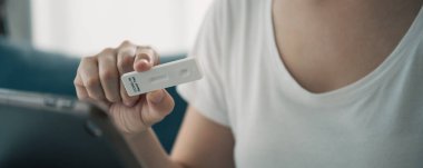 SARS 2019-nCoV COVID-19 Coronavirus antijen hızlı test kiti kullanan, hijyen koruyucu yüz maskesi takan Asyalı genç bir kadın - evde tarım test kiti ve video aracılığıyla doktorla test sonuçlarına danışmak.