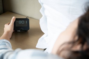 Tembel Asyalı genç bir kadın, yatağın yanındaki masada uyanan dijital çalar saati uyutmaya çalışıyor. Kapatın, kadın sabah 8: 00 'de uyanır. Yatak odasında yatakta uyumaya çalışan bir kadın..