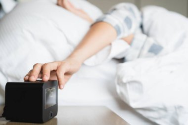 Tembel Asyalı genç bir kadın, yatağın yanındaki masada uyanan dijital çalar saati uyutmaya çalışıyor. Kapatın, kadın sabah 8: 00 'de uyanır. Yatak odasında yatakta uyumaya çalışan bir kadın..