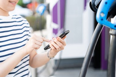 Şarj istasyonu, Bev veya elektrikli araç şarj istasyonu ile akıllı telefon bağlantısı kurmak için her türlü şarj uygulamasını kullanan mutlu Asyalı genç kadın. EV istasyonunda yakıt ikmali yapan kadın..