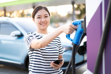 EV şarj istasyonunda DC-CCS tip 2 EV şarj konnektörü tutan mutlu Asyalı kadın, bir EV-elektrikli araç konnektörü hazırlayan kadın bir aracı şarj etmek için.