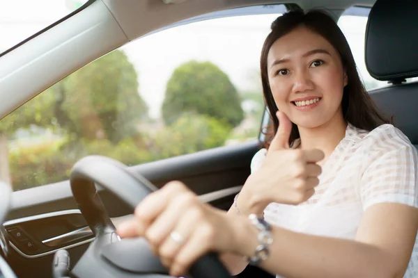 Happy Asian woman driving the electric vehicle - EV car smiling and showing her thumb up to the camera, happy and safety driving concept. Woman driving modern EV car and smiles to camera.