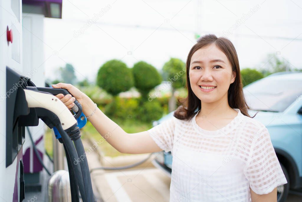 Happy Asian woman holding a DC - CCS type 2 EV charging connector at EV charging station, woman preparing an EV - electric vehicle charging connector for recharge a vehicle.