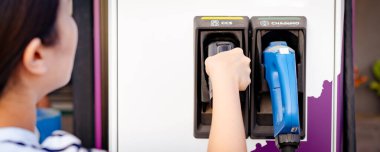 DC 'yi tutan tanınmayan Asyalı kadın - EV şarj istasyonunda CCS tip 2 EV konnektörü, bir EV-elektrikli araç hazırlayan kadın bir aracı şarj etmek için konnektör.