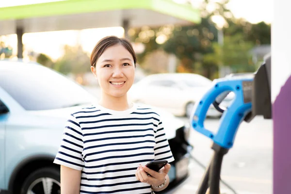 아시아인젊은 여성은 스마트폰 스마트폰에 사용하여 Bev 자동차 스테이션에서 전지를 용하는 — 스톡 사진