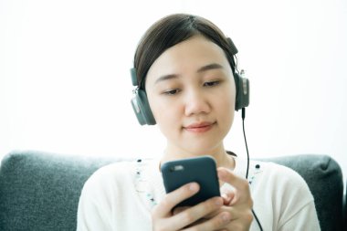 Asyalı genç bir kadın oturma odasında dinleniyor ve telli kulaklık kullanarak akıllı telefondan derin derin düşüncelere dalmaktan zevk alıyor. Çalışma hayatı yaşam tarzı kavramını dengeler.