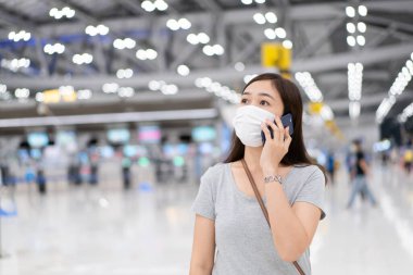 Güzel Asyalı, koruyucu maskeli genç bir kadın havaalanında akıllı telefondan konuşuyor. Coronavirüs 'te seyahat ederken maske takan kadın - COVID-19 salgını.