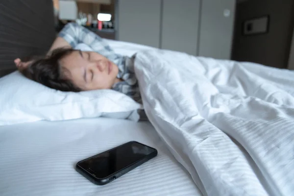 Lazy Asian young woman trying to snooze awaking alarm clock in smartphone close up, woman wake up late in morning up to 8.00 am. Tried woman sleeping on the bed in bedroom.