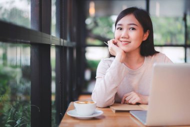 Çalışan Asyalı güzel bir kadın kahvecide ya da kafede içki içiyor. Bilgisayarına ya da bilgisayarına yazı yazan neşeli bir internet blogcusu ve yakın mesafeden kahve içiyor. Modern yaşam tarzı.