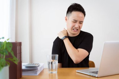 Siyah tişörtlü Asyalı genç adam çok çalışmaktan ya da çok çalışmaktan sağlık sorunu yaşıyor. Gençlerde kritik sağlık sorunu ve ofis sendromu hastalığı. Yeni normal yaşam tarzlarıyla evden çalışmak.