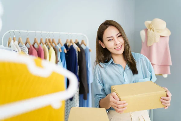Happy Asian young woman showing a cardboard parcel box to camera, online selling or ecommerce concept. Female online seller sells a clothes via online marketplace platform.