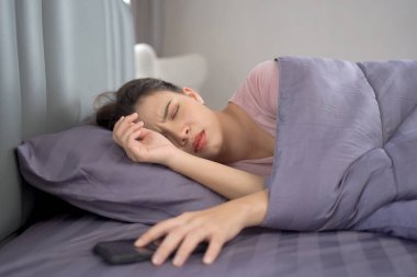Güzel Asyalı genç bir kadın sabahları yatakta uyuyor ve akıllı telefondan çalar saati kapatmaya ya da uyutmaya çalışıyor. Tembel genç kadın, sabah geç kalkıyor..
