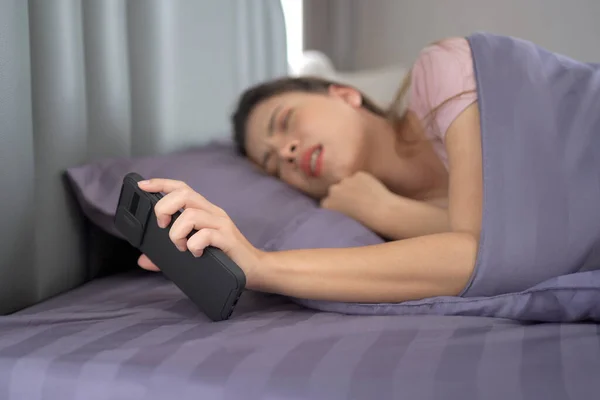 Beautiful Asian young woman sleeping on the bed in morning and trying to turning off or snoozing the alarm clock on smartphone. Lazy young woman get up late in morning.