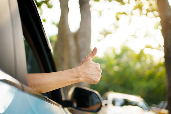 Unrecognizable Asian woman driver showing a thumb up outside the vehicle while driving a car. Happy woman driver or passenger showing her thumb up during in the car. Safety trip and freedom concept.
