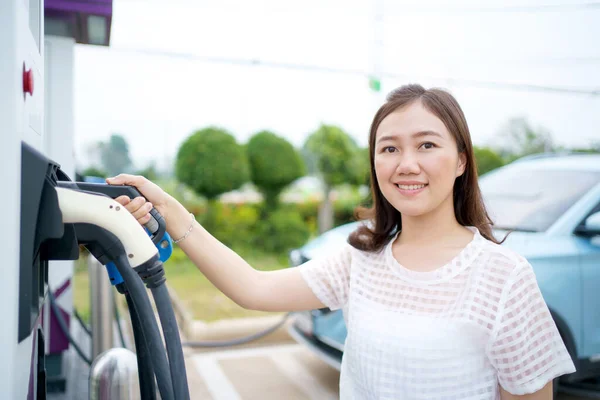 Happy Asian woman holding a DC - CCS type 2 EV charging connector at EV charging station, woman preparing an EV - electric vehicle charging connector for recharge a vehicle.