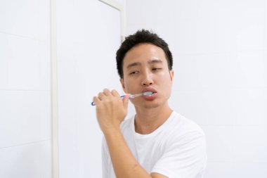 Beyaz gömlekli Asyalı genç bir adam sabah yataktan kalktıktan sonra aynanın karşısında dişini fırçalıyor, ağız sağlığı ve sağlık hizmetleri. Diş sağlığı konsepti.
