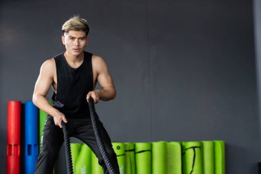 Güçlü Asyalı sporcu, kapalı bir salonda kopyalanmış halat egzersizi yapmak için hazır bekliyor. Güçlü genç sporcu vücut geliştirme konsepti geliştiriyor, kas geliştiriyor..