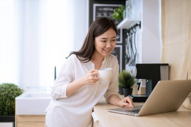 Neşeli Asyalı kadın sabahları bir fincan kahve içip internette sosyal medya aramaktan hoşlanıyor. İş kadını laptopta e-postayı kontrol ediyor ve sabahları kahve içiyor.