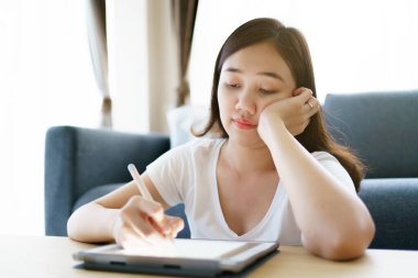 Happy Asian young woman relaxing in the living room and watching a contents and chatting with her friends and drawing an illustration on tablet. Woman enjoy watching video and drawing on tablet.