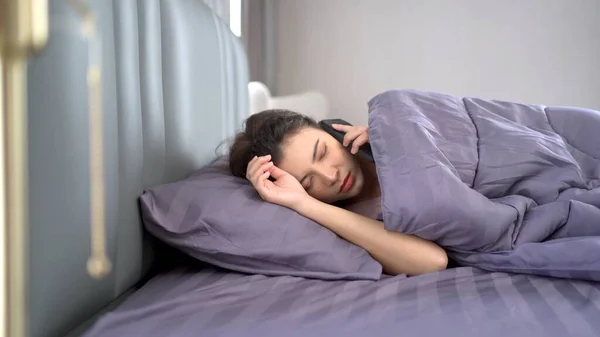 Beautiful Asian young woman sleeping on the bed in morning and trying to turning off or snoozing the alarm clock on smartphone. Lazy young woman get up late in morning.