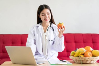 Hastanedeki ofisinde çalışan mutlu Asyalı beslenme uzmanı. Ofiste dizüstü bilgisayar kullanarak bir hastayla sohbet etme konusunda uzman. Profesyonel beslenme danışmanı portresi.