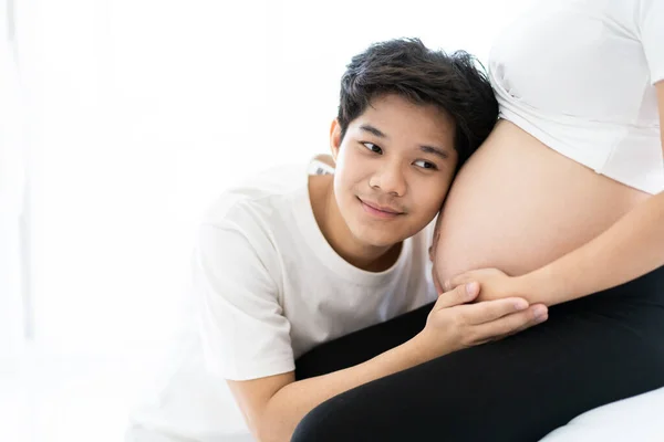 Lovely Asian husband gently listening on a belly of his pregnant wife while laying on the bed together. Father feels excited while listening on his wife\'s stomach. Concept of happy family.