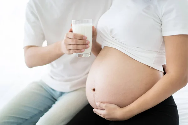 Lovely Asian couple taking care together in their house, husband taking care his pregnant wife by giving a fresh milk. Wellness and health care in pregnancy woman and unborn child in tummy.