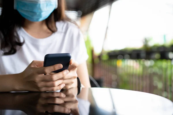 Woman wear a face mask while relaxing at cafe and restaurant, woman texting a message on a smartphone close up with copy space. Asian young adult using protective face mask during stay outdoor.