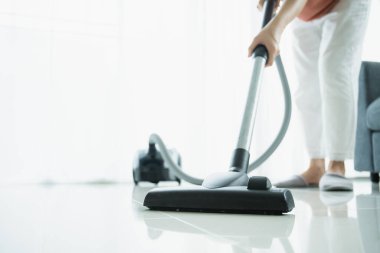 Asyalı genç temizlikçi, oturma odasındaki kirli zemini temizlemek için elektrikli süpürge kullanıyor. Kopyalama aletiyle kapatıyor. Ev hanımı oturma odasında yerleri ve kanepeleri süpürüyor..