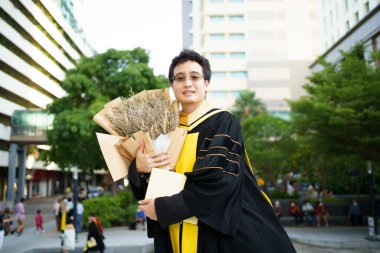 Üniversite mezuniyet töreninde elinde güzel bir buket çiçek tutan mutlu Asyalı öğrenci. Elbise giymiş yüksek lisans öğrencisi fotoğraf için bir buket çiçek gösteriyor..