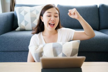 Happy Asian young woman relaxing in the living room and watching a contents and chatting with her friends and drawing an illustration on tablet. Woman enjoy watching video and drawing on tablet.