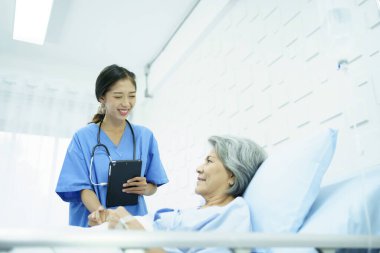 Hastanedeki tıp yatağında dinlenen yaşlı Asyalı kadın hasta doktorla konuşuyor. Uzman doktor, Asyalı yaşlı bir hastayı ziyaret edip onunla konuşuyor..