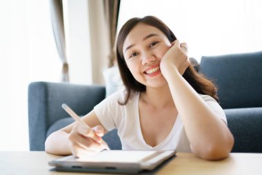 Happy Asian young woman relaxing in the living room and watching a contents and chatting with her friends and drawing an illustration on tablet. Woman enjoy watching video and drawing on tablet.