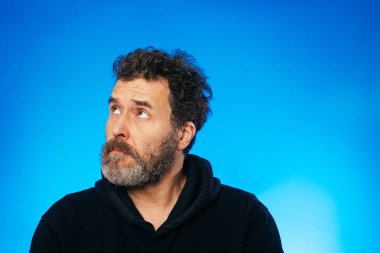 Kuşkucu orta yaşlı sakallı adam yukarı bakıyor, jestler yapıyor ve mavi arka planda stüdyo resimlerinde duygularını gösteriyor.