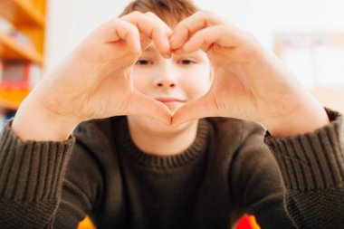 Ten year old school boy showing heart shape hands in home video schooling situation, close up looking into wide angle camera