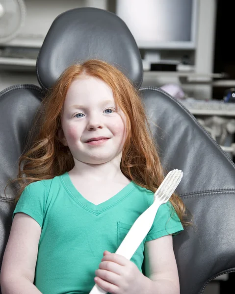 Niña sentada en una silla de dentista sosteniendo un cepillo de dientes gigante — Foto de Stock