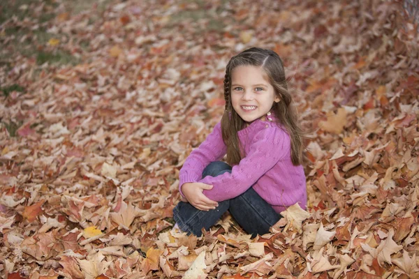Sorridente bambina caucasica seduta nelle foglie autunnali Foto Stock Royalty Free
