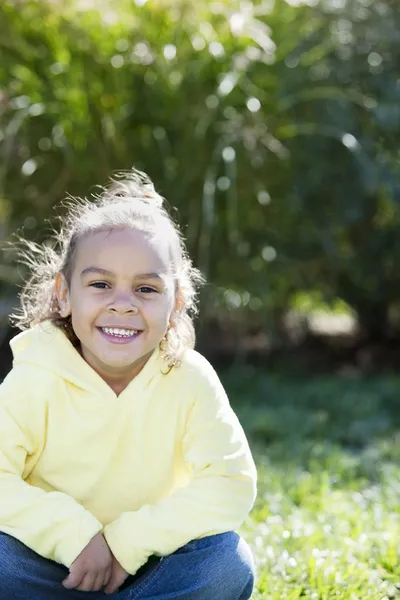 Real: Leende blandad ras liten flicka sitter gärna i en solig park — Stockfoto