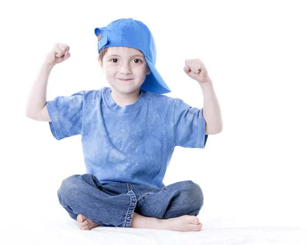 Sonriente niño caucásico sentado con piernas cruzadas flexionando sus músculos —  Fotos de Stock