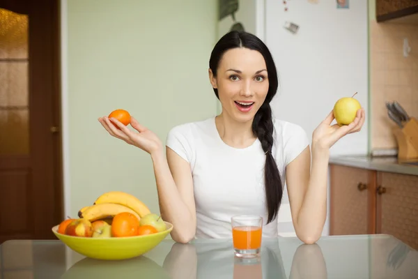 Brunette girl holding fruits sitting in the kitchen — Stock Photo, Image