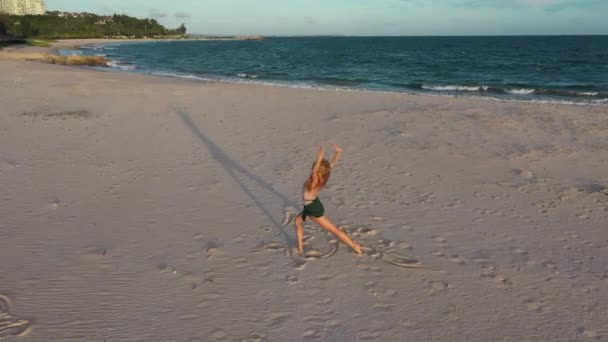 若い美しい少女バレリーナは ゆっくりとした動きで黄金の夕日の海の近くのビーチで踊っています 感動的な女性 魅力的な女優の笑顔 海岸のダンサーの近くでドローンが飛ぶ — ストック動画