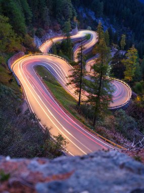 İsviçre, Maloja 'nın dağ geçidi. Ormanda birçok virajlı bir yol. Araba ışıklarının bulanıklığı. Akşamları manzara. Seyahat için geniş çözünürlüklü fotoğraf