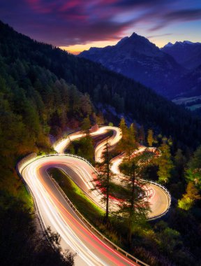 İsviçre, Maloja 'nın dağ geçidi. Ormanda birçok virajlı bir yol. Araba ışıklarının bulanıklığı. Akşamları manzara. Seyahat için geniş çözünürlüklü fotoğraf