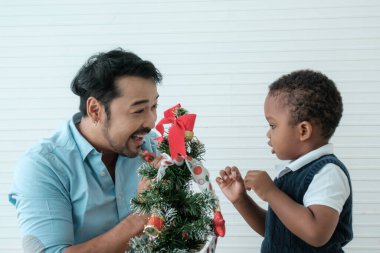 Daddy and son enjoy using times to decrorate christmas tree and take a selfie with mobile at home.