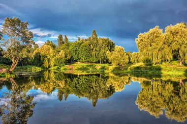 Nehir kıyısında harika bir gün batımı, ağaçlar su ve üzerlerindeki fırtına bulutları yansıtılır.