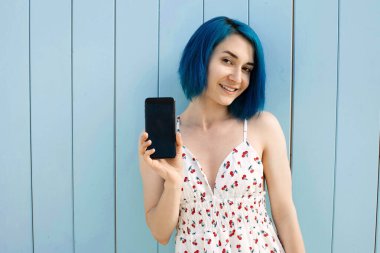 Cyan duvarının yanında dikilirken boş ekran cep telefonu gösteren gülümseyen beyaz bir kadının portresini kapat.