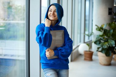 A caucasian female freelancer holdnig a laptop and looking in a window in a co-working.