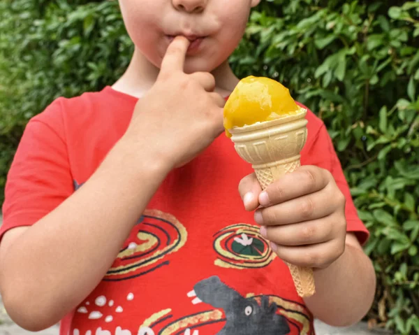 Çocuk elinde sarı bir dondurma tutuyor ve parmağını yalıyor.