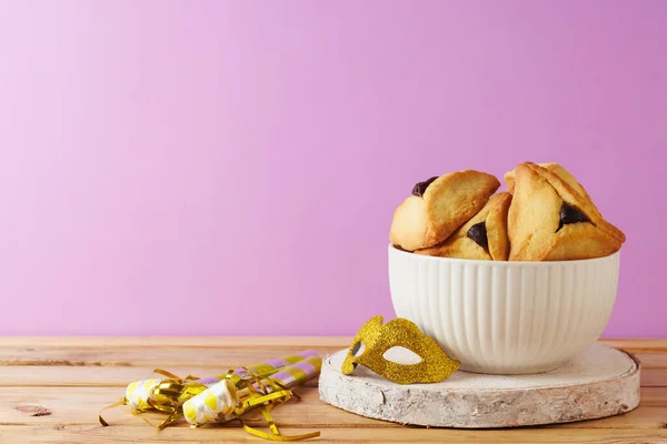 ユダヤ人の休日紫色の背景の上に木製のテーブルの上にHamantaschenクッキーとカーニバルマスクとプリムコンセプト — ストック写真
